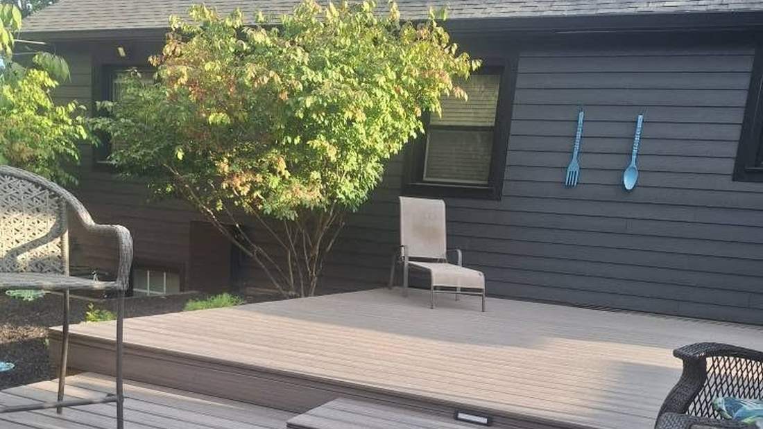 A refinished composite deck with railings and patio chairs, against the fresh grey-stained siding of a house.